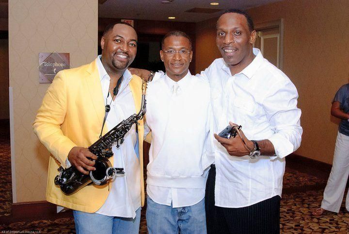 Three men posing indoors, one holding a saxophone and another holding a microphone.