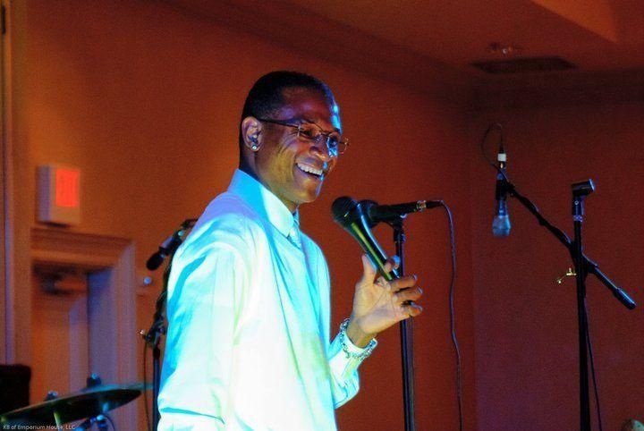 Speaker at a microphone on a warmly lit stage, wearing a white shirt and gesturing while speaking.