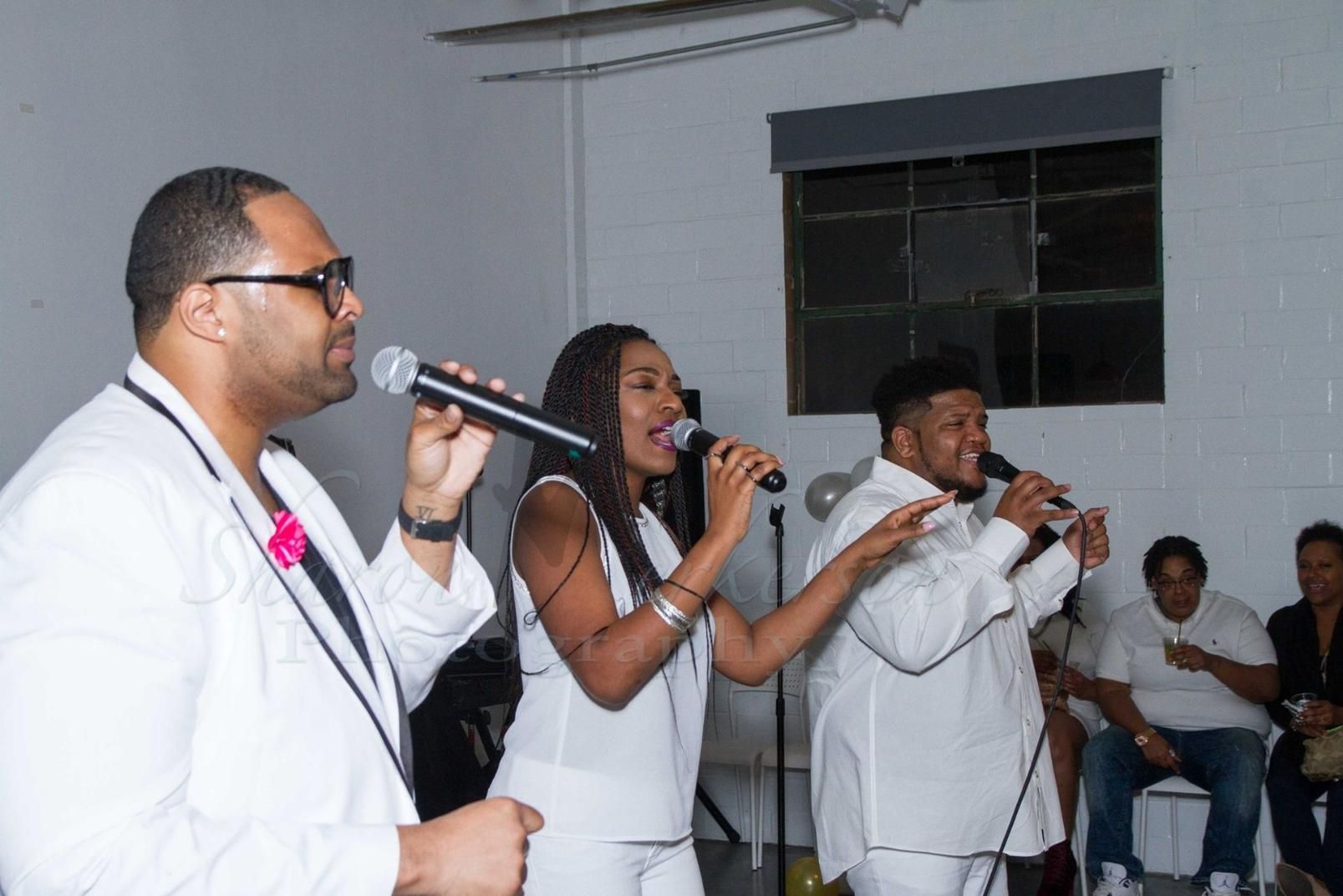 Three singers in white perform with microphones on a small indoor stage.