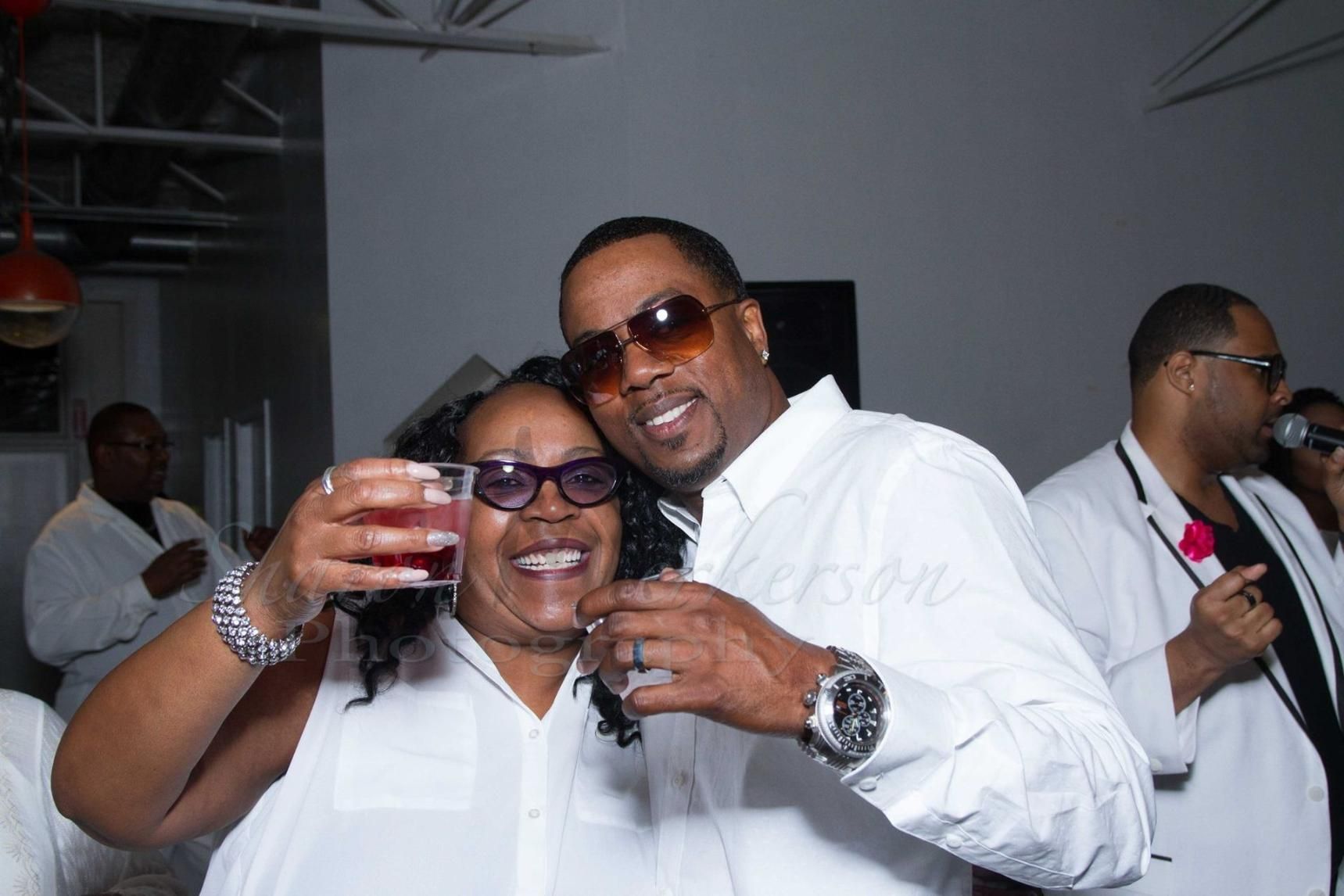 Two people in white shirts toast drinks and smile at an indoor party.