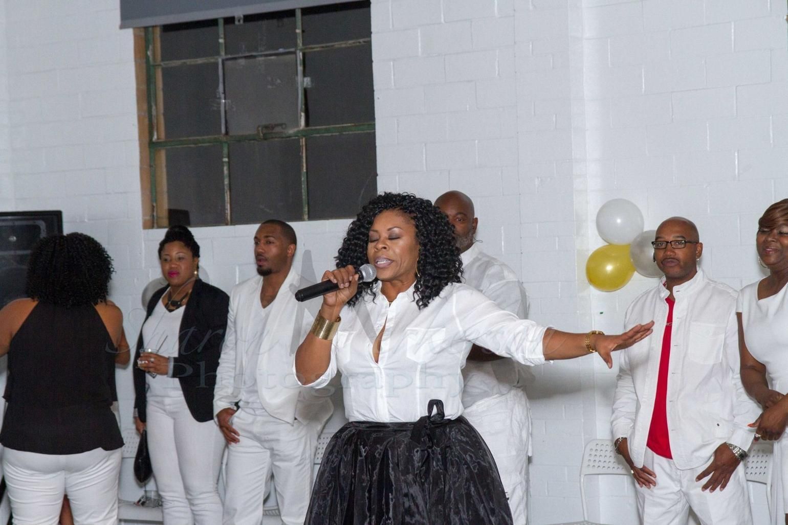 Woman singing into a microphone at an indoor event, wearing a white shirt and black skirt, with people in the background