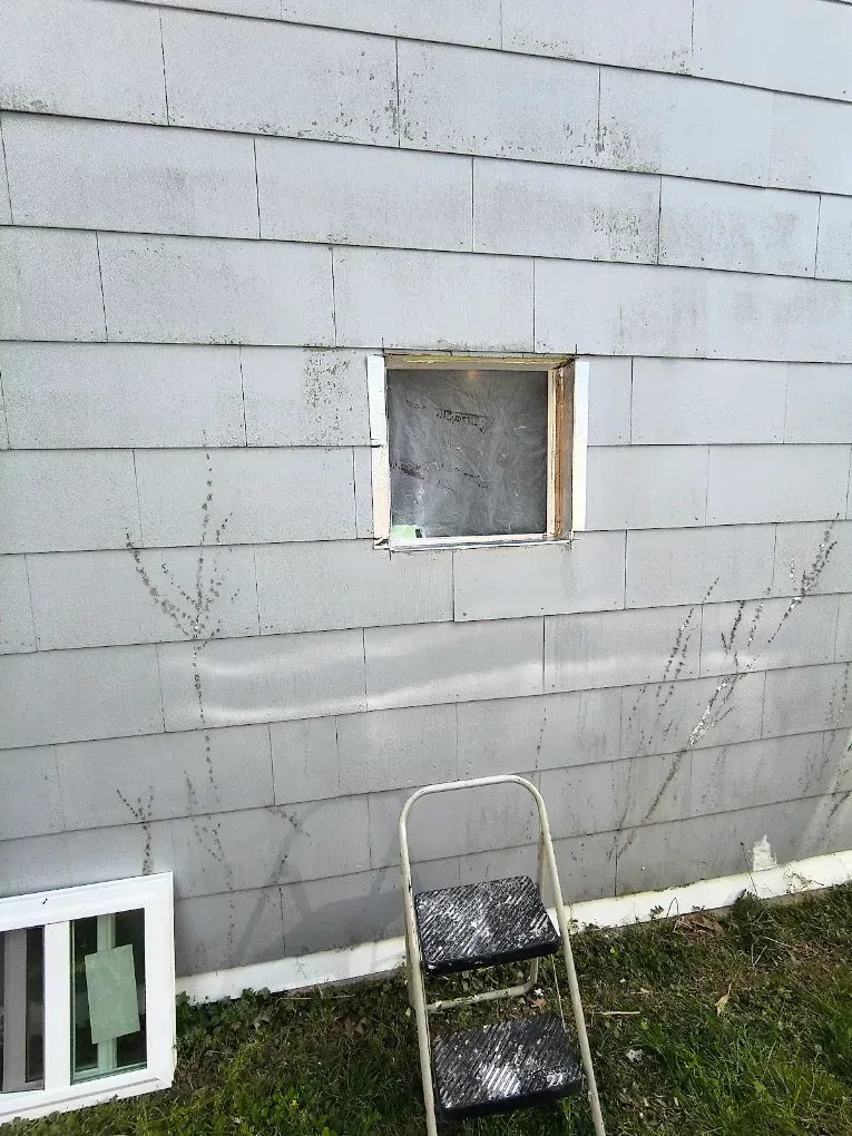 Gray cinder block wall with a small window, leaning metal step stool, and a vent on the grass below.