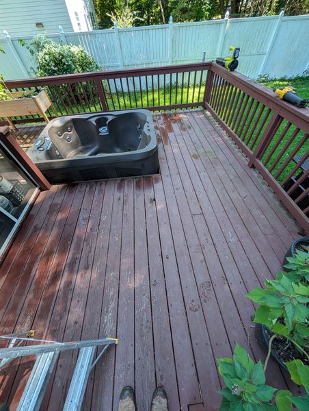 Wooden deck with a hot tub and railing, bordered by plants and a white fence