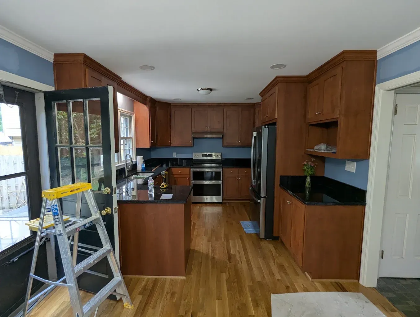 Kitchen interior with wood cabinets, black countertops, stainless appliances, and a step ladder near the window.