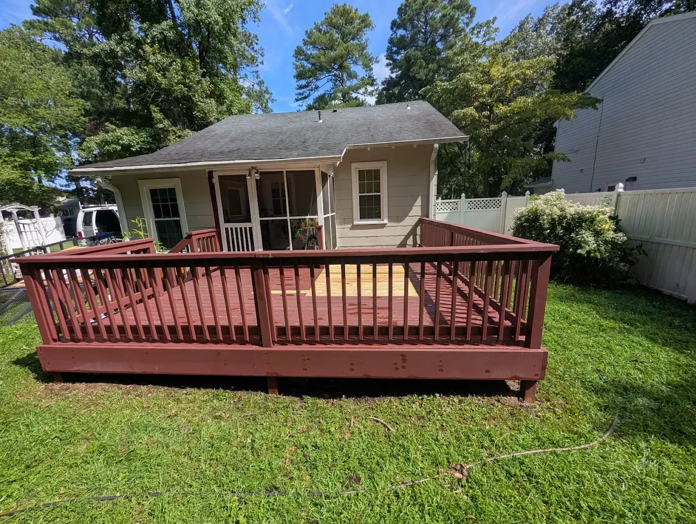 Small beige house with a red wooden deck and railing in a grassy backyard under trees