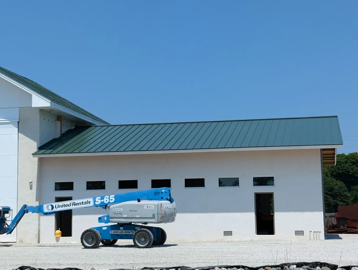 Blue boom lift parked beside a white building with green roof and garage doors under a clear sky