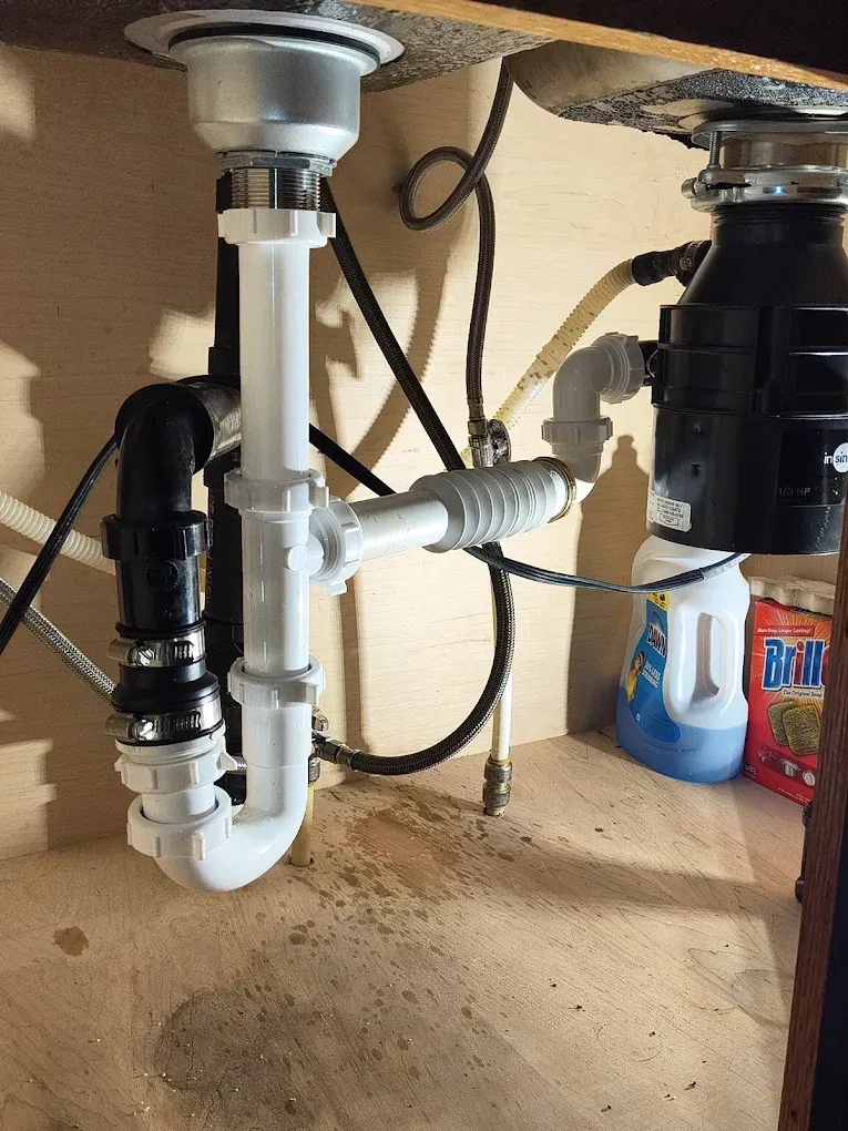 Under-sink plumbing with white and black drain pipes and a garbage disposal in a cabinet