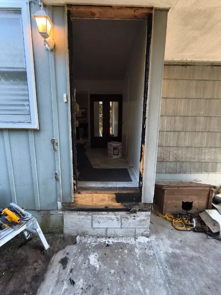 Front doorway of a house with open door, lit porch light, concrete steps, and scattered tools on the patio.