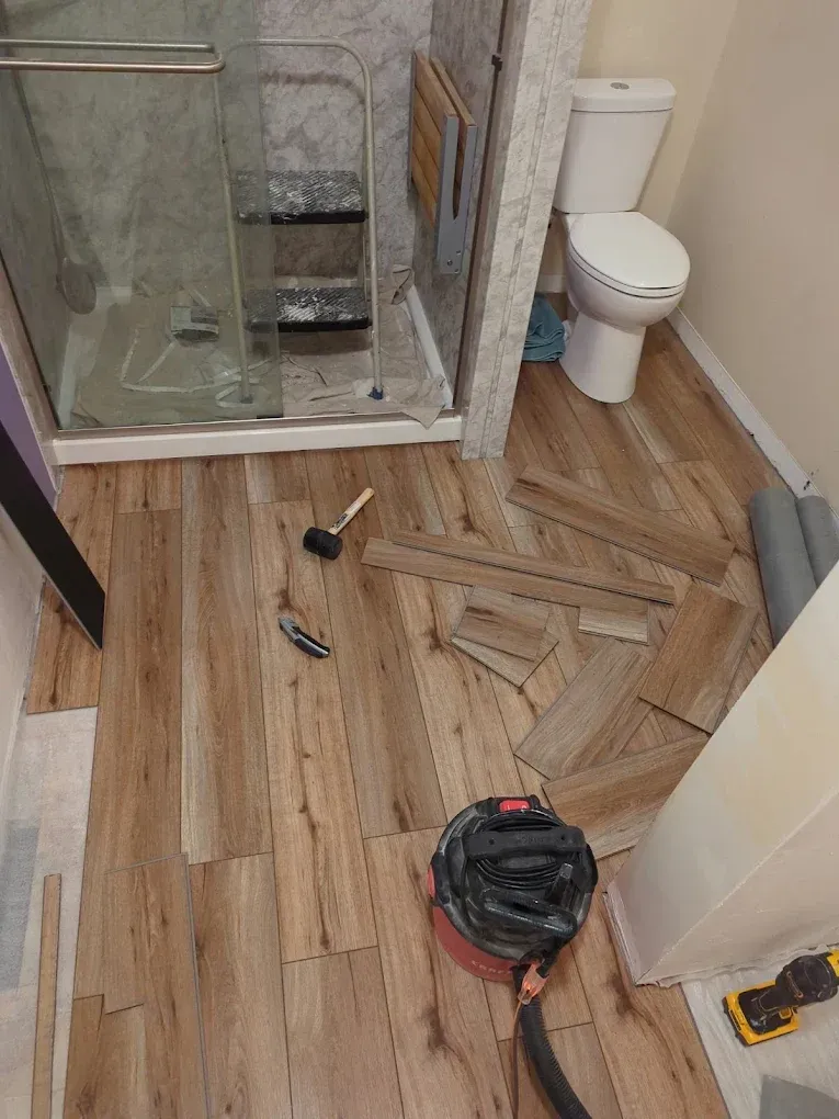 Bathroom under renovation with broken floor tiles, tools, and debris near the shower and toilet.