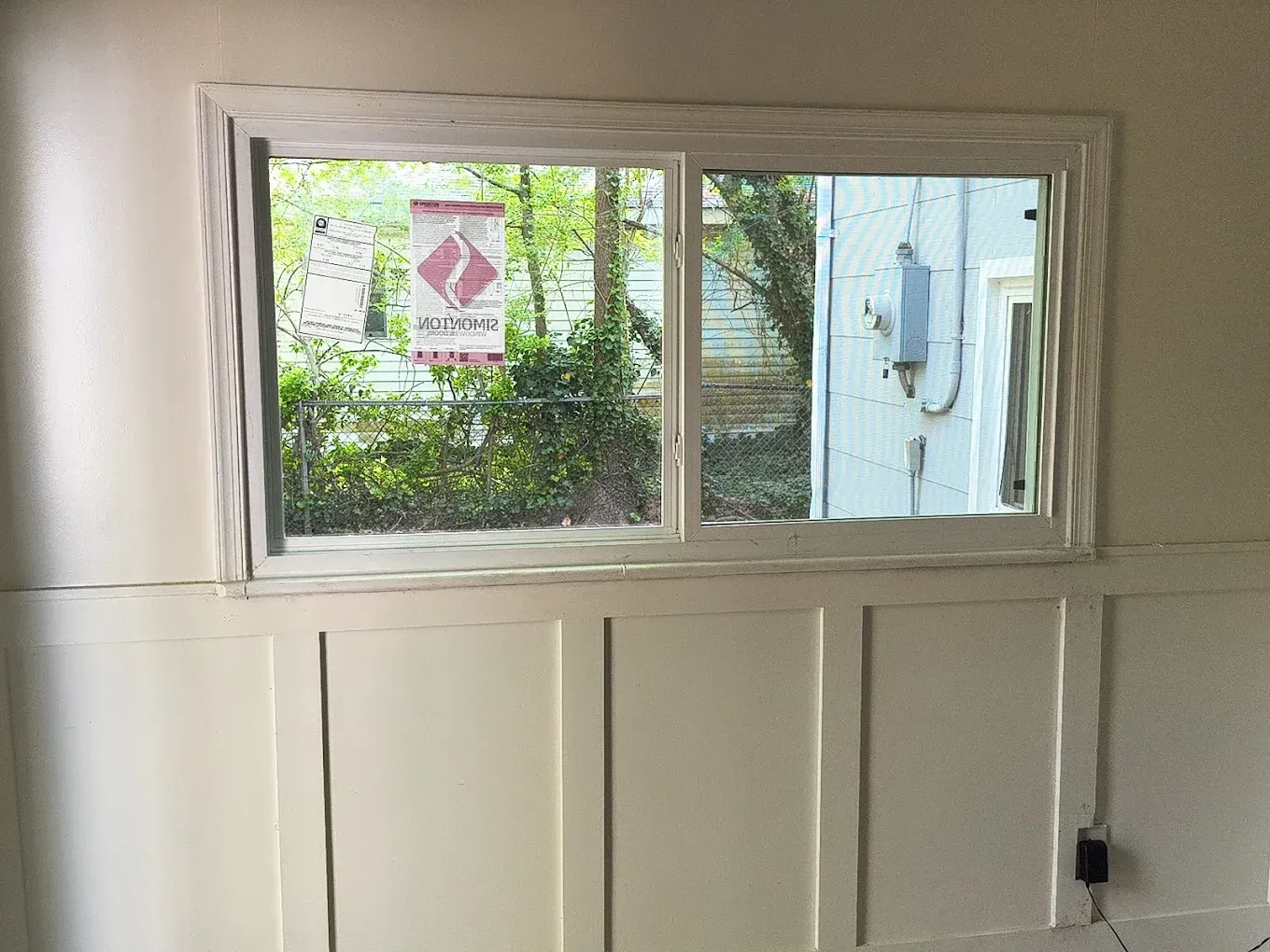 Small window above white paneled wall with a view of greenery and a neighboring building outside.