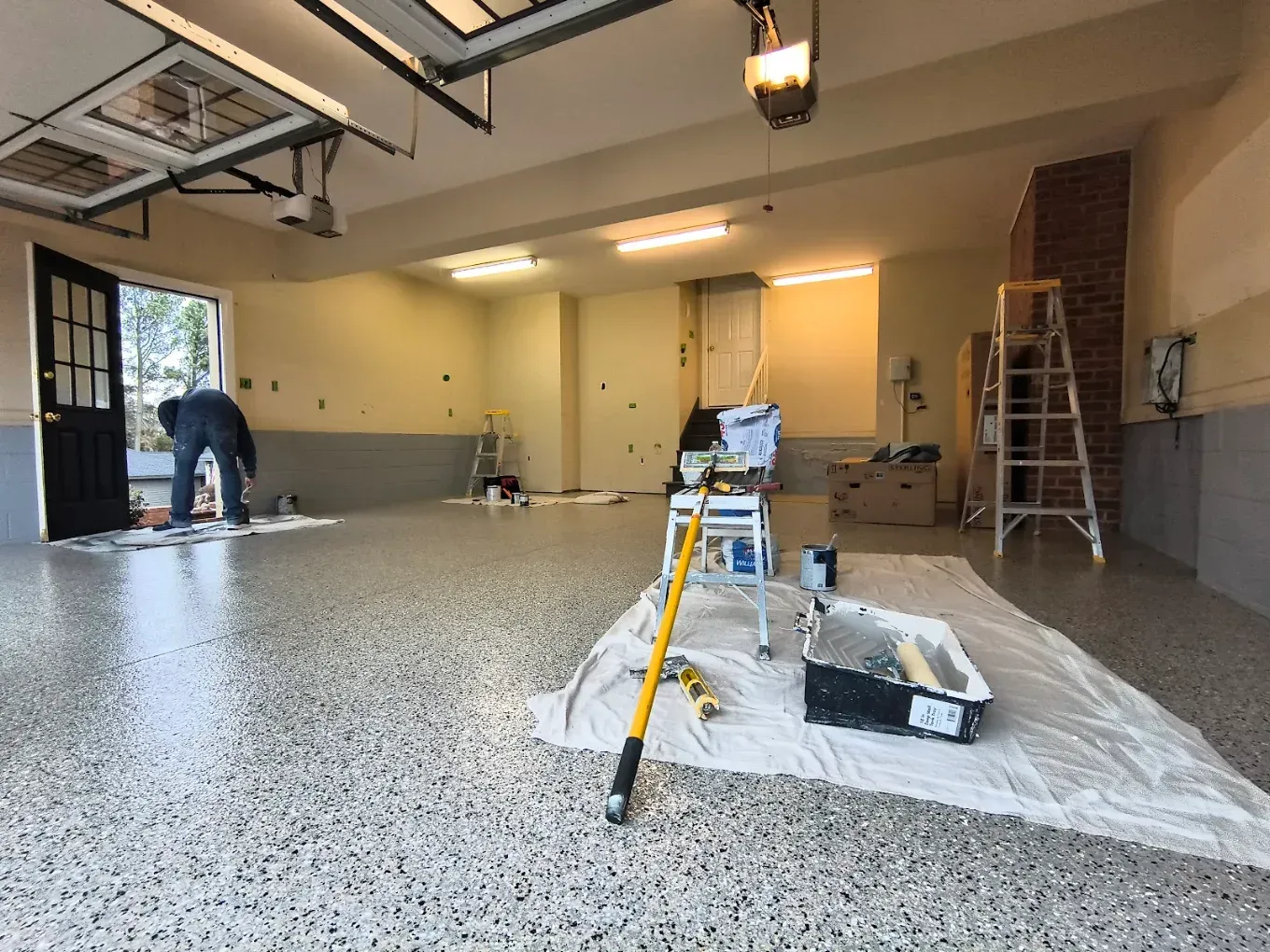 Garage interior being coated with speckled epoxy floor; workers and tools visible, ladder at right.