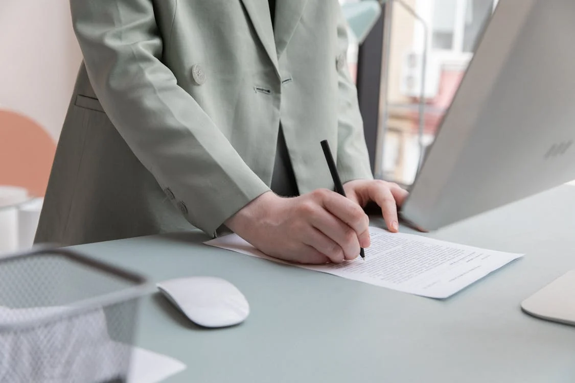 Person writing on a notepad beside a laptop at a desk in a bright office