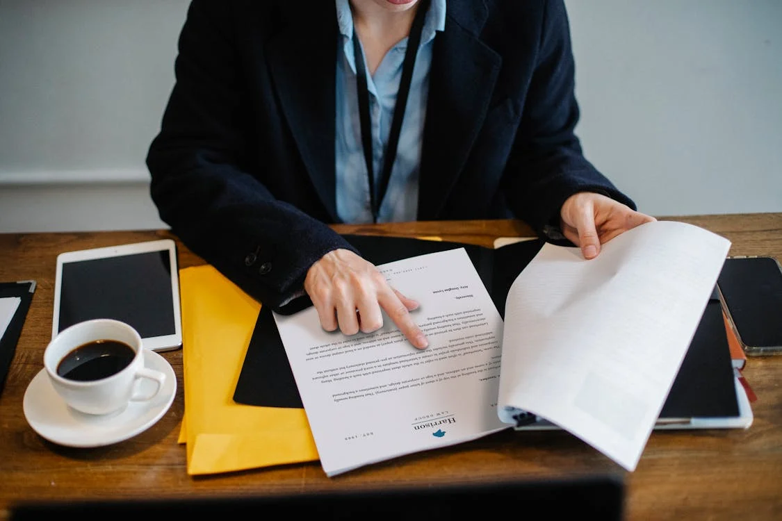 Person flipping through papers at a desk with a coffee cup, tablet, and yellow folder