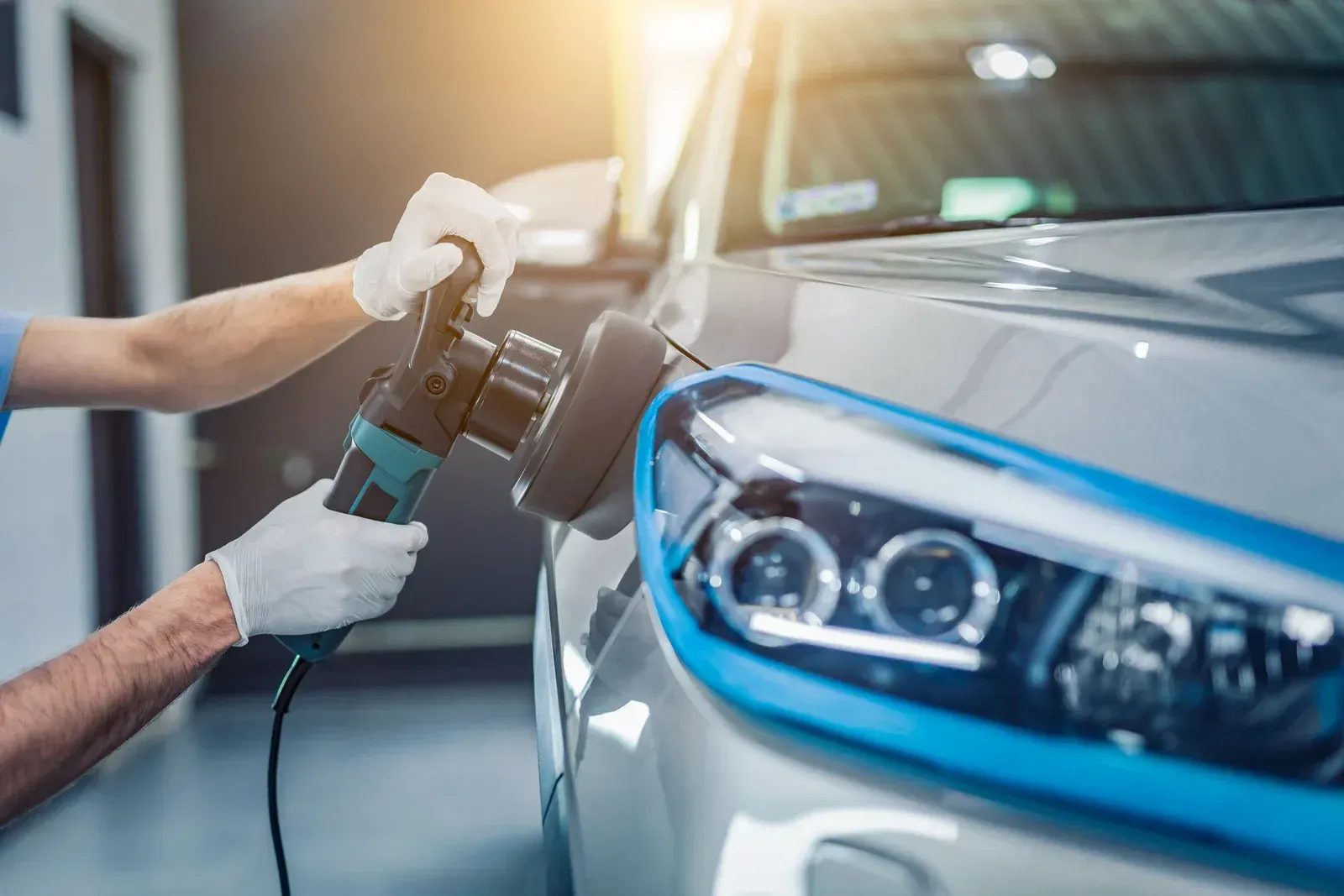 Person polishing a silver car's headlight with a power buffer, wearing gloves, in a garage.