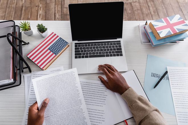 Vista dall'alto di una scrivania con computer portatile, documenti, libri e mani che esaminano dei documenti con bandiere degli Stati Uniti e del Regno Unito.