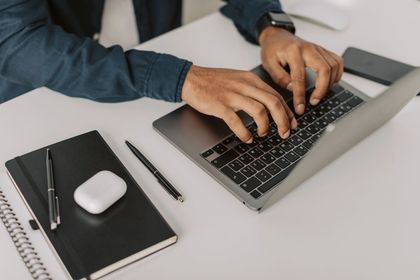 Mani che digitano su un laptop accanto a un quaderno, una penna e un mouse wireless su una scrivania bianca.