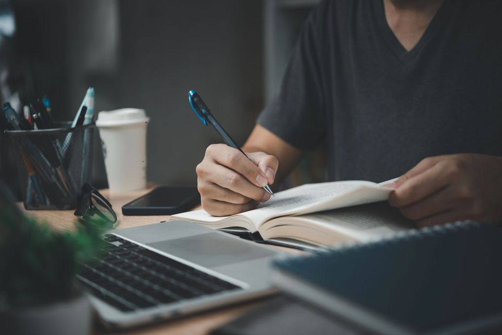 Una persona scrive su un quaderno accanto a un computer portatile, con una tazza di caffè sulla scrivania.