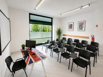 Aula vuota con file di sedie nere, banchi, pareti bianche e una grande finestra con vegetazione all'esterno.