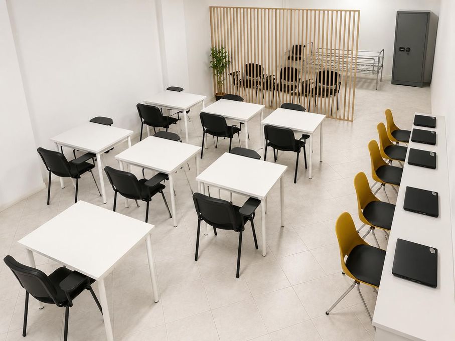 Aula moderna e vuota con tavoli bianchi, sedie nere e una fila di sedili gialli lungo la parete.