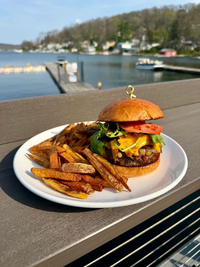 Burger and fries on a white plate by the water with a blurred marina in the background