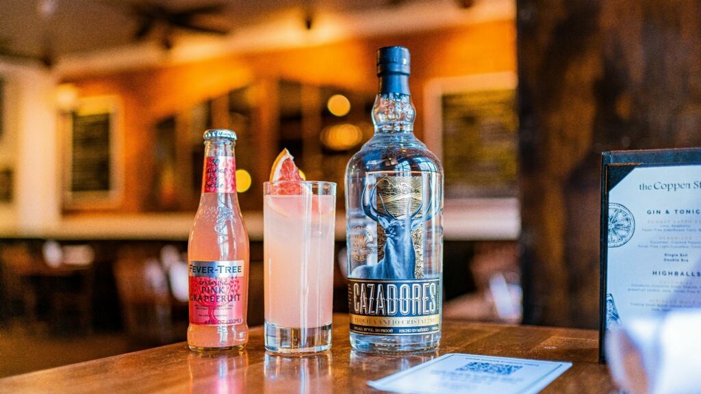 Bottle and cocktail on a bar table, with a menu in the foreground and warm pub lighting.