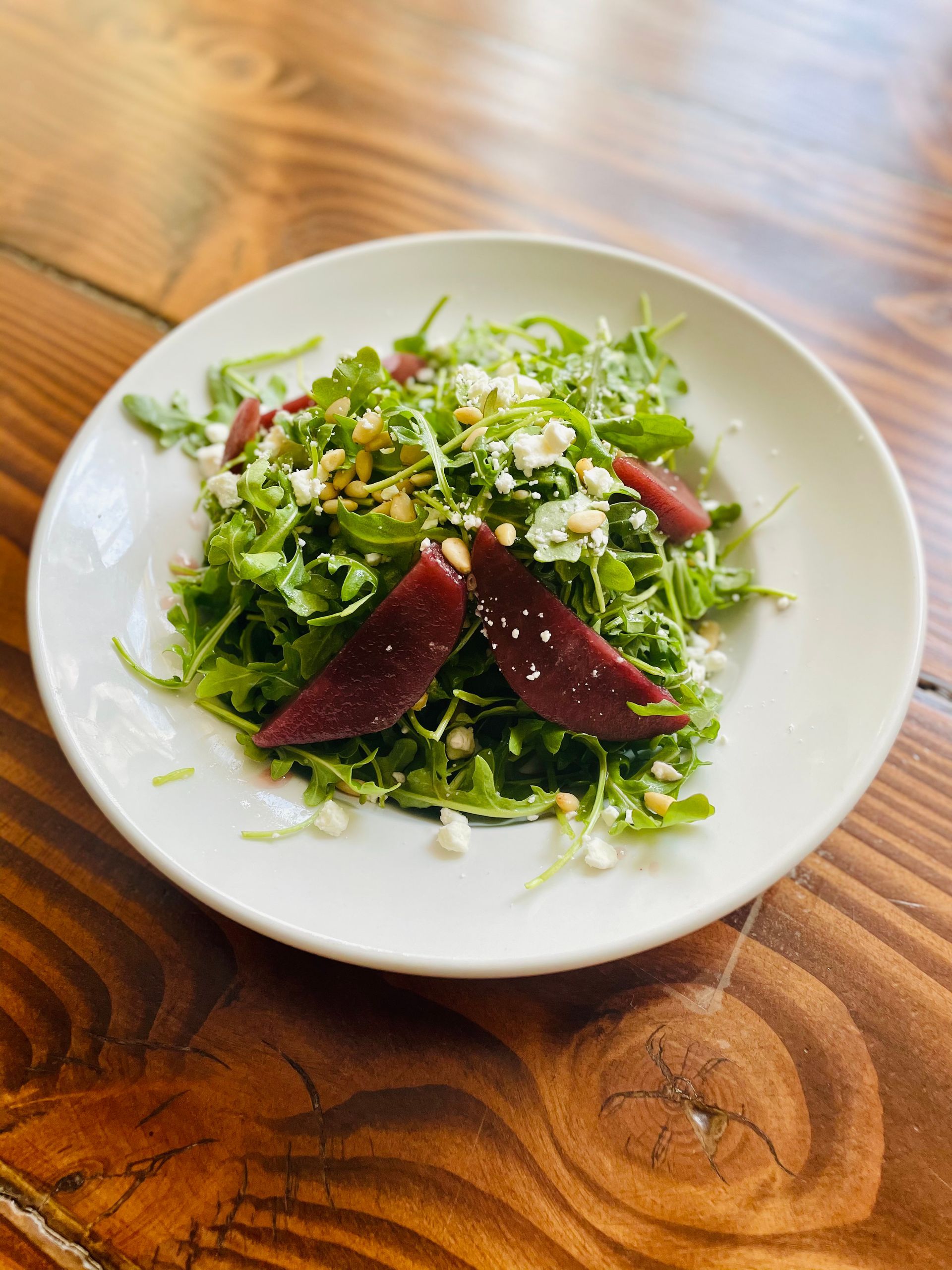 Mixed green salad with shaved cheese and beet slices on a white plate atop a wooden table