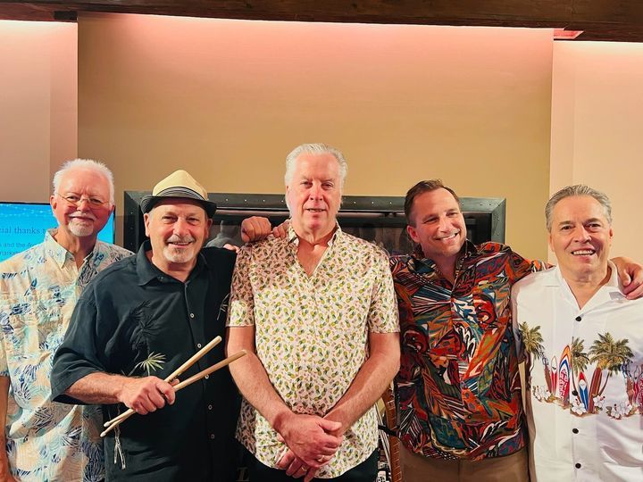 Five men smiling indoors, one holding drumsticks, in casual patterned shirts at a gathering