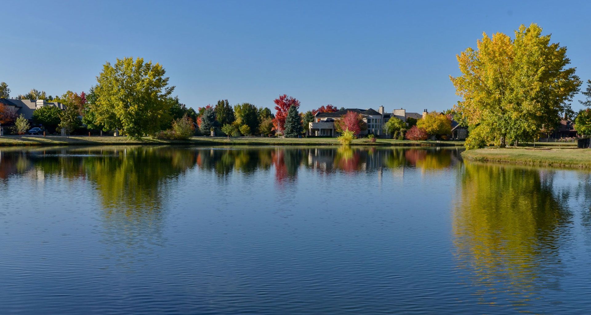 Calm lake reflecting autumn trees and houses under a clear blue sky