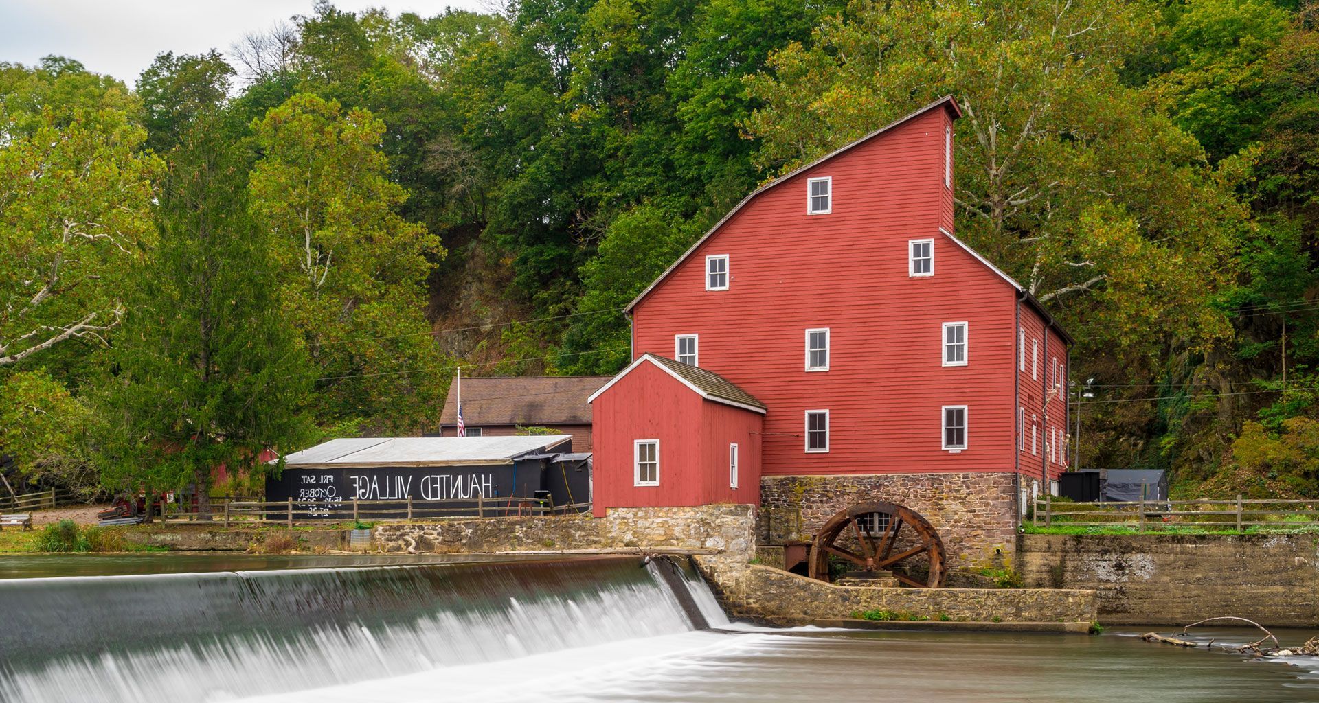 Red mill by a river dam with a small bridge and green trees in the background
