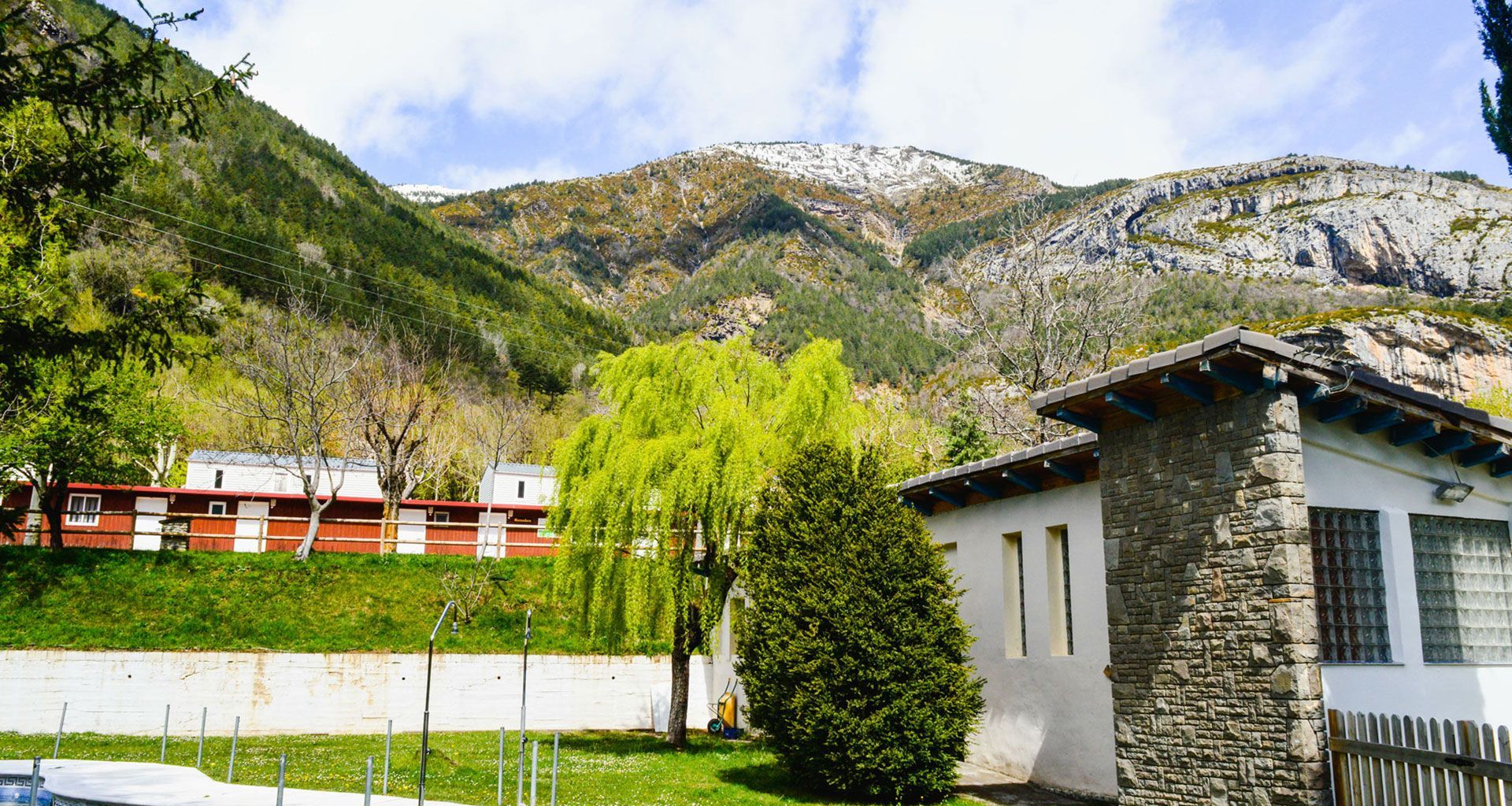 Mountain village with white houses, a stone building, green trees, and rocky hills under a cloudy sky