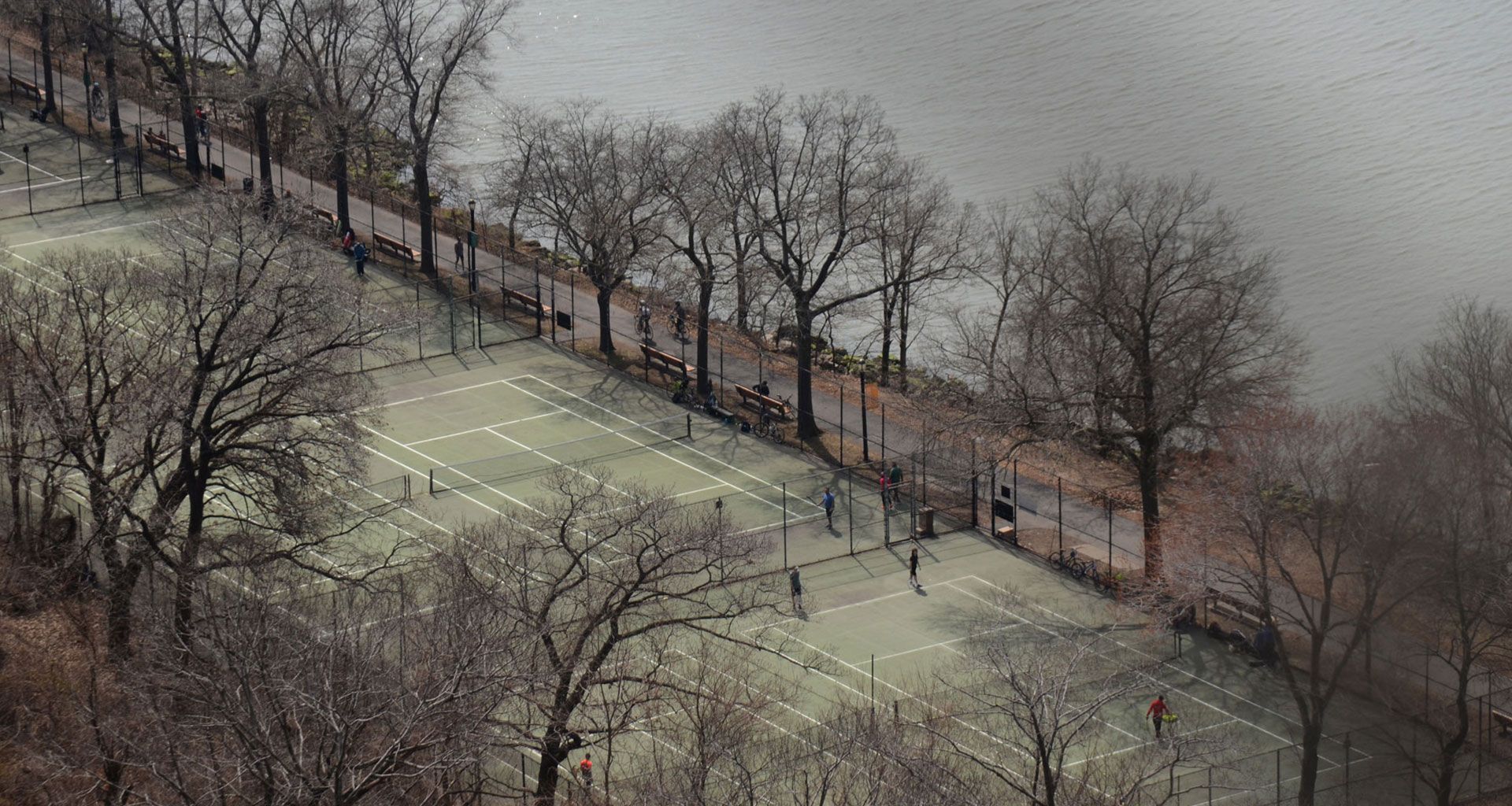 Snow-covered tennis courts by a riverside, with bare trees and a few people walking along the path.