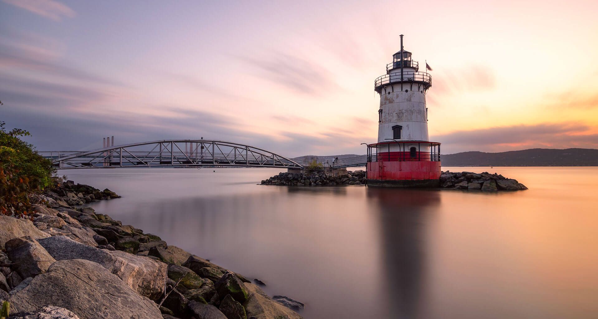 Lighthouse on a rocky breakwater at sunset with calm water and a bridge in the background