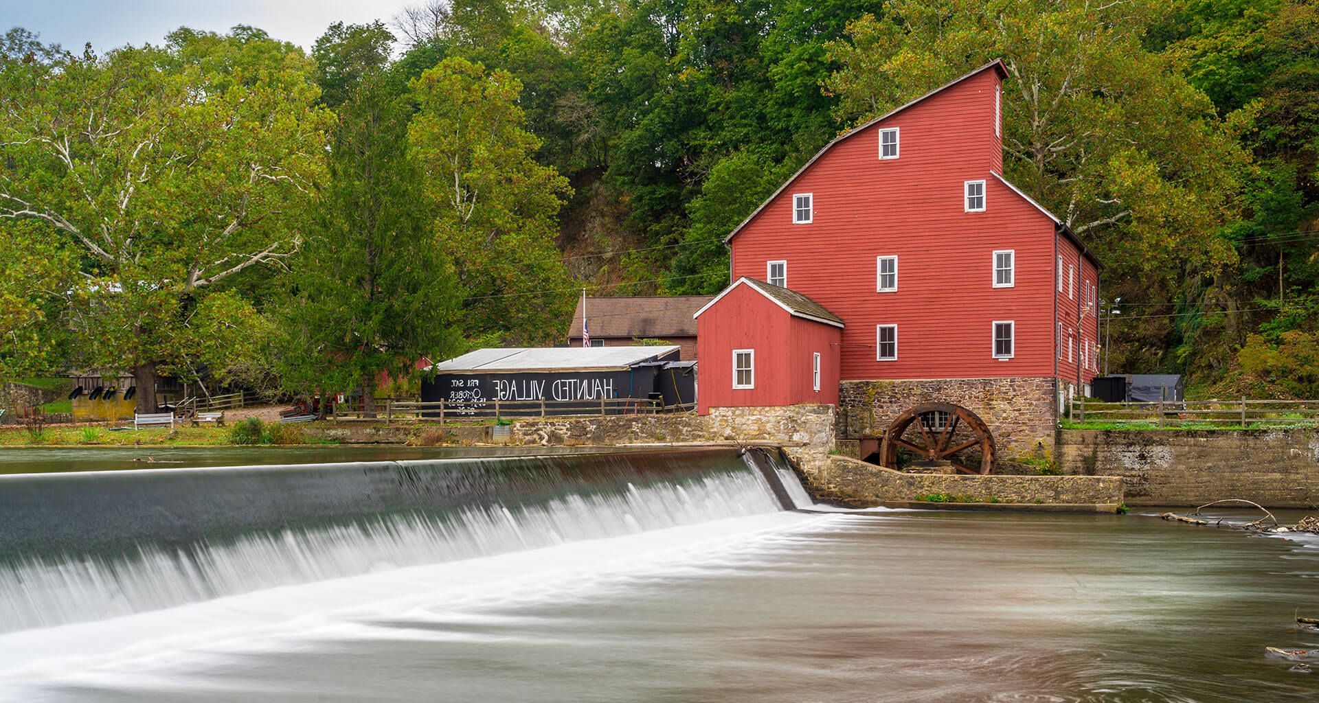 Red mill beside a small waterfall on a river, with trees and a cloudy sky.
