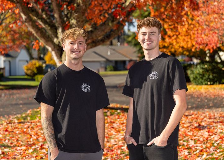 Two people standing on a leafy suburban street, smiling in black T-shirts and jeans.