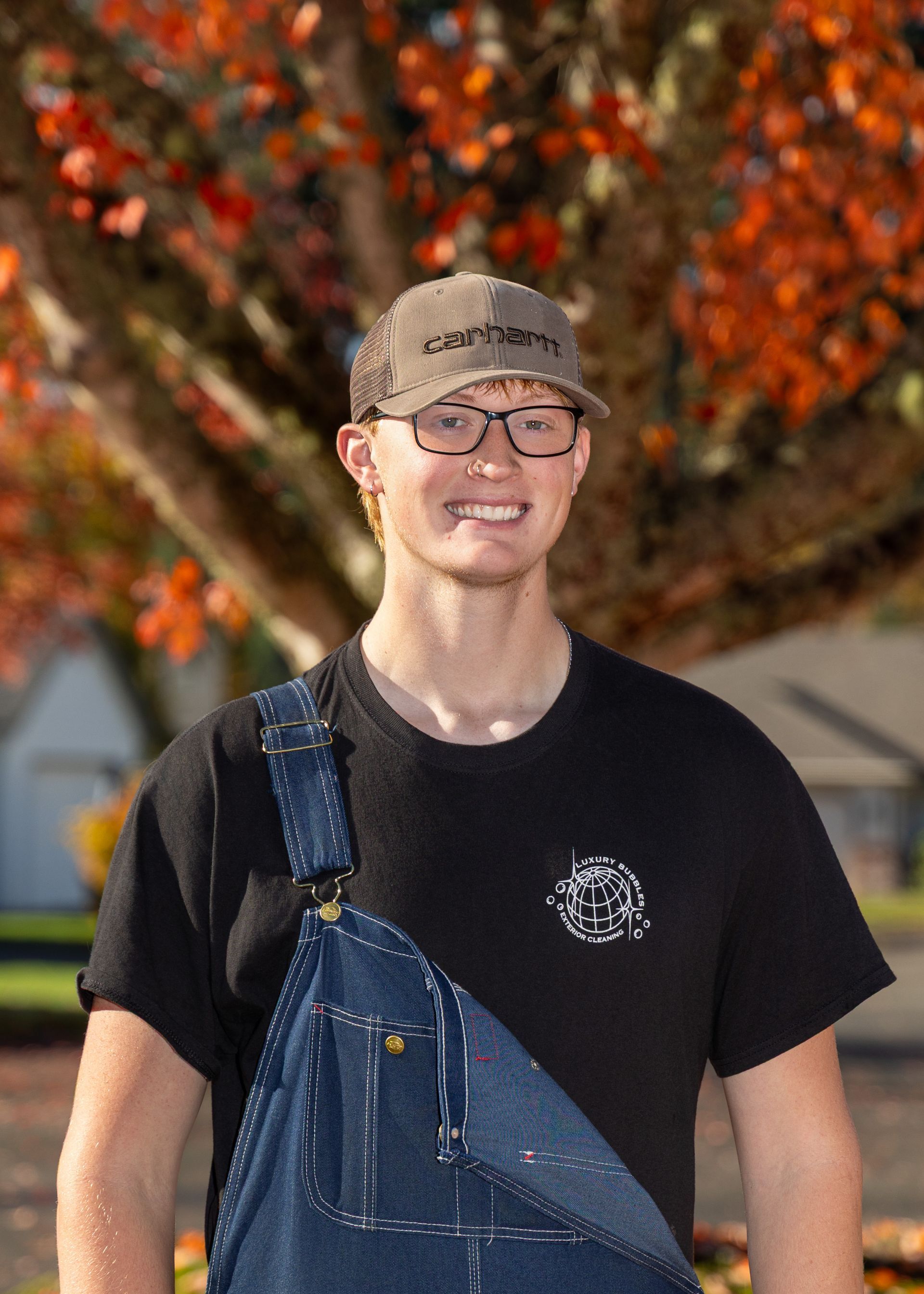 Smiling person in glasses and cap wearing denim overalls, standing outdoors with autumn leaves behind them