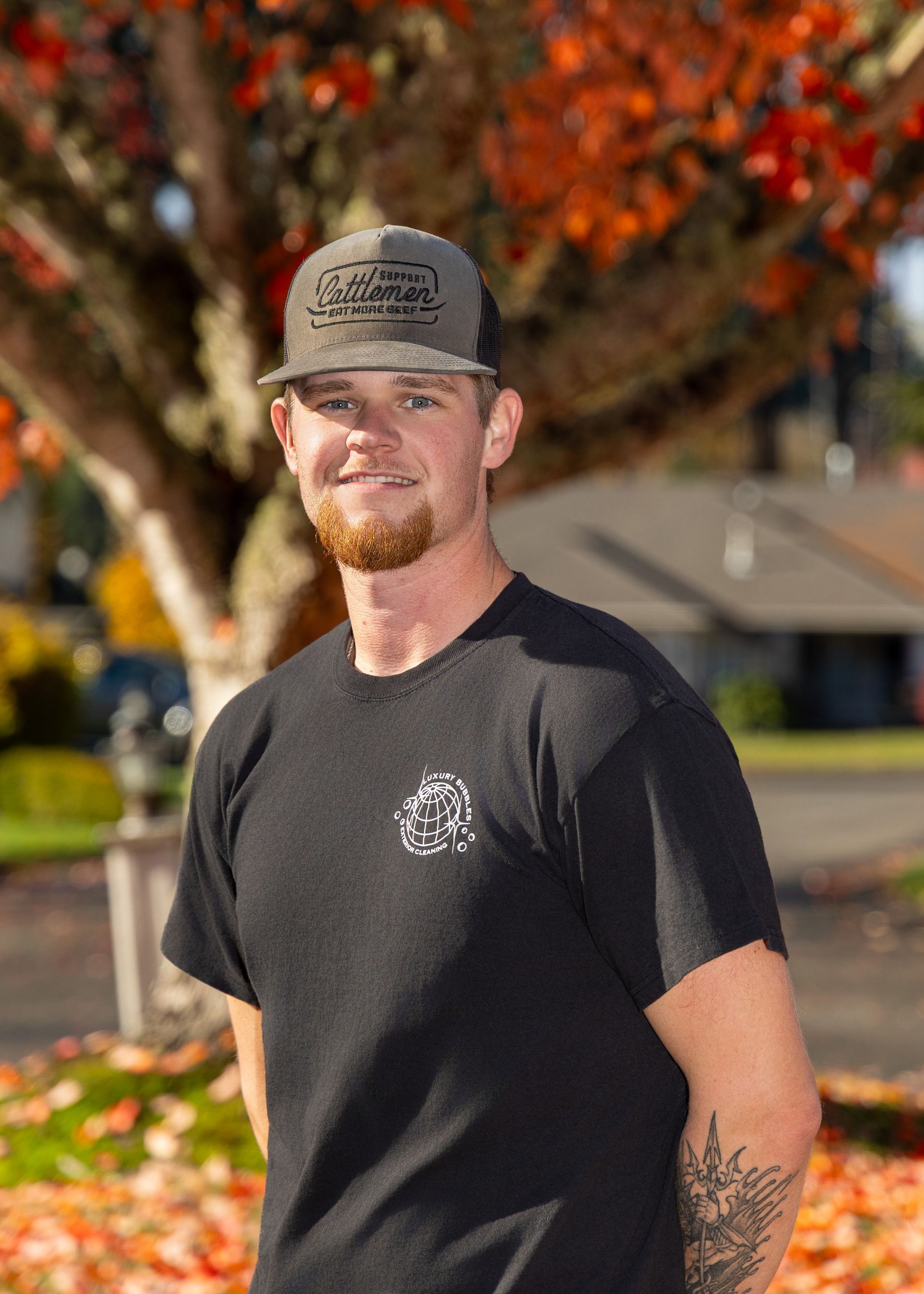 Man in a black T-shirt and cap standing outdoors with autumn trees in the background