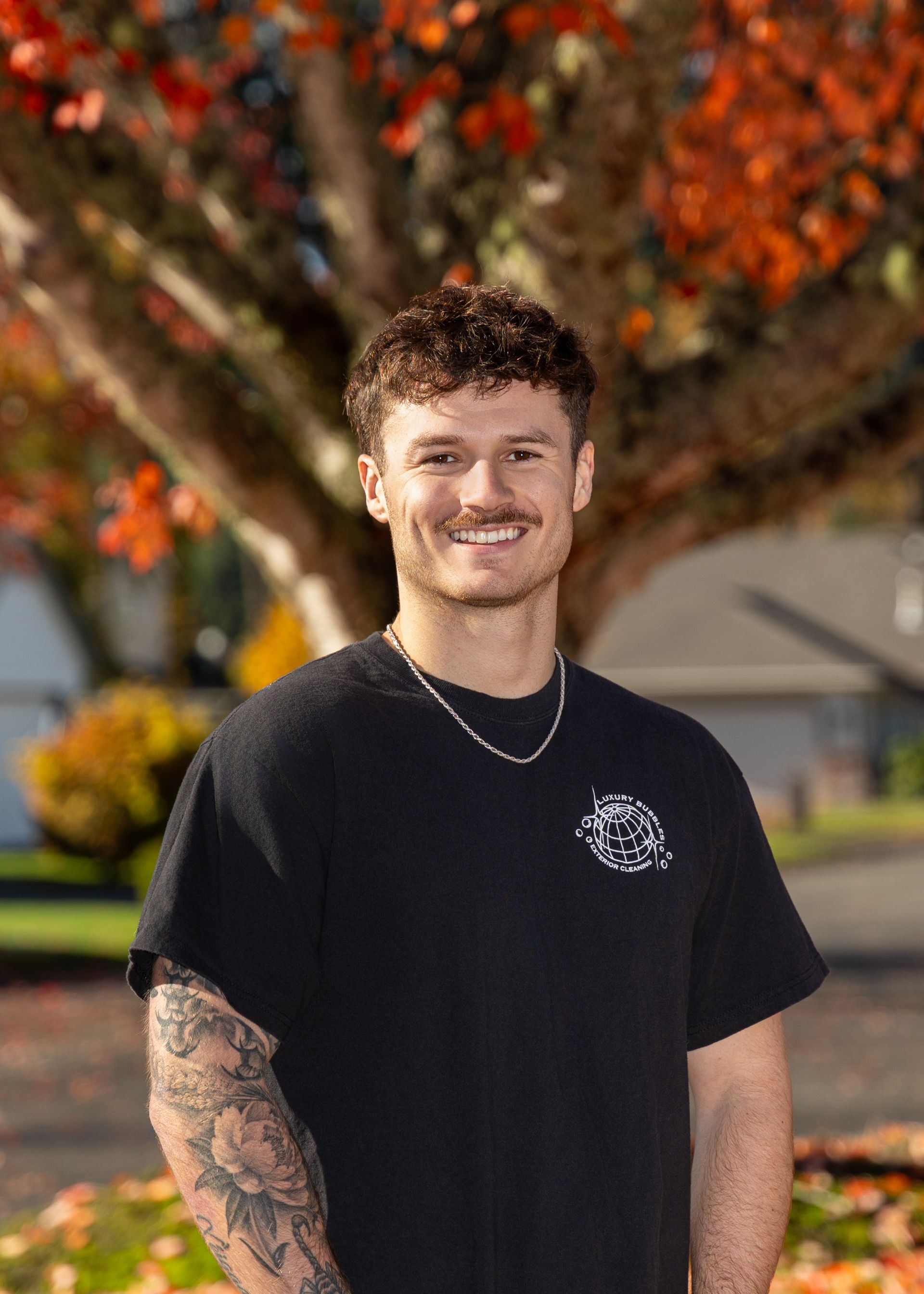 Smiling person in a black T-shirt standing outdoors with autumn leaves in the background
