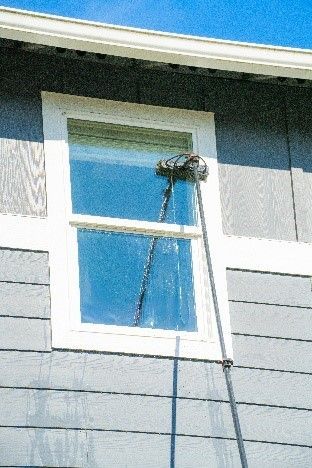 Window being washed from the outside with a long-handled brush, house siding visible.