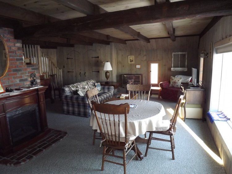 Rustic living room with plaid sofa, round table, fireplace, wood-paneled walls, and bright window light