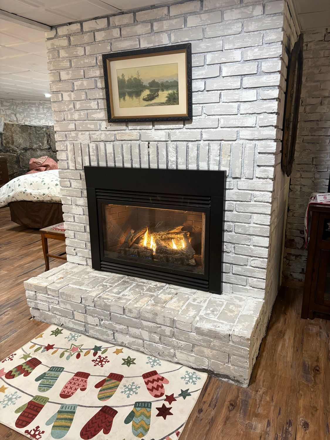 White brick fireplace with lit fire, framed artwork above, and a holiday rug on hardwood floor.