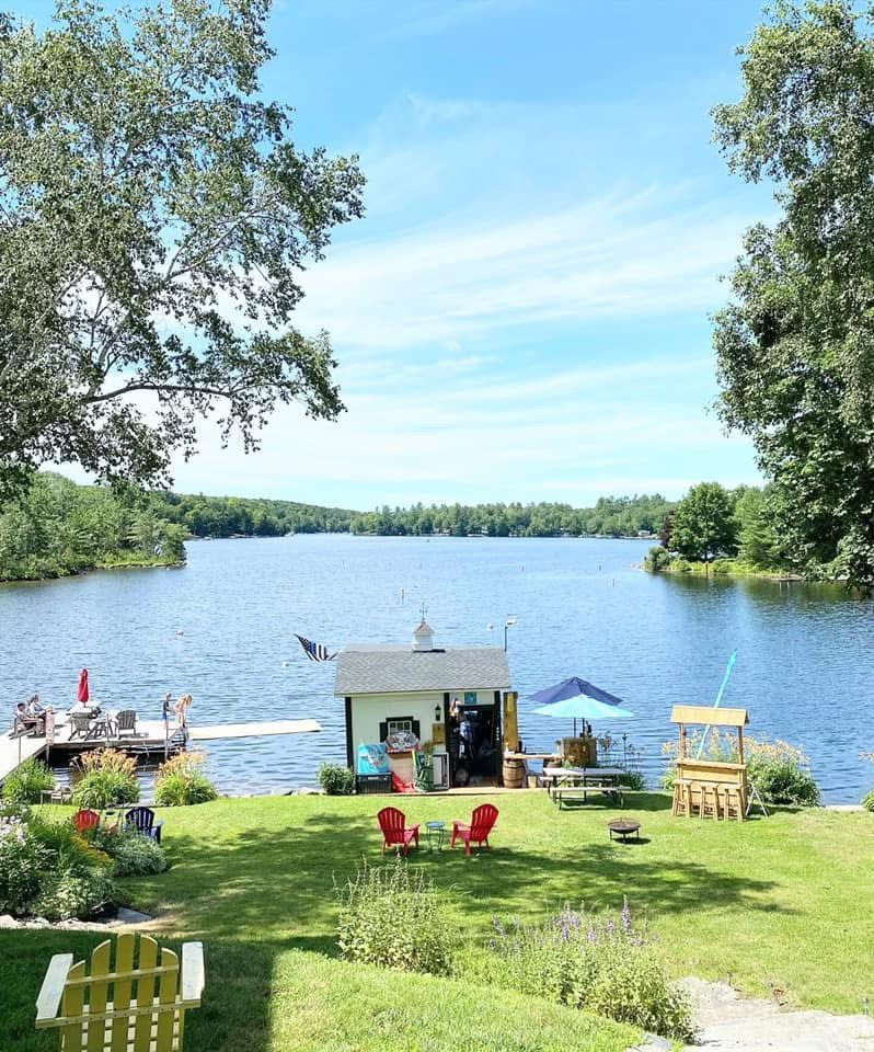 Lakeside lawn with dock, chairs, umbrella, and a small boathouse under a blue sky