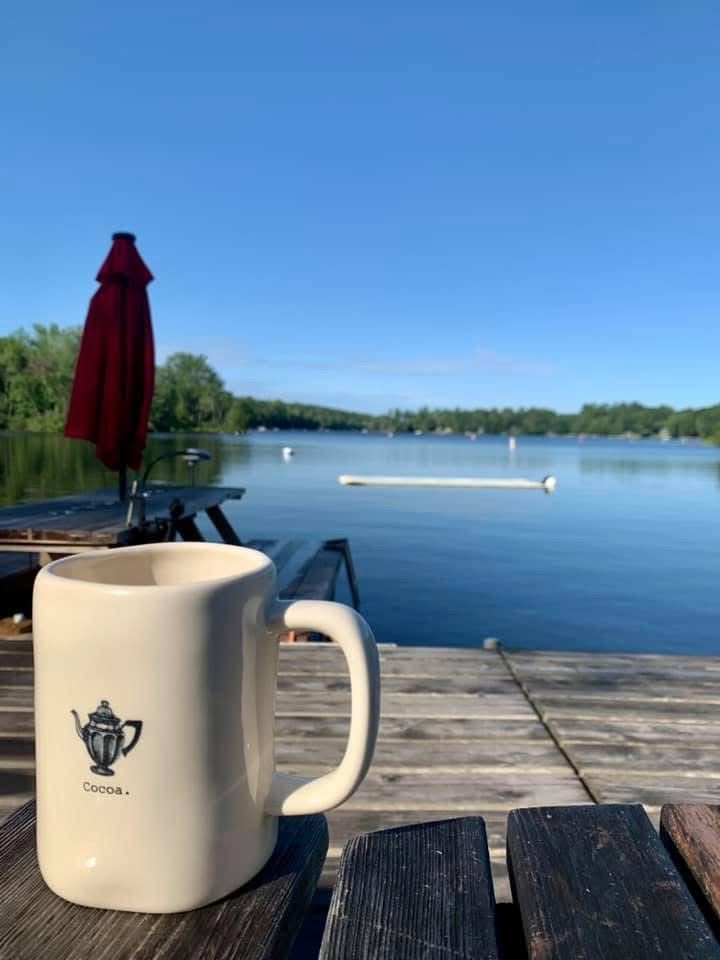 White mug on a wooden dock beside a red umbrella, overlooking a calm lake under a clear blue sky.