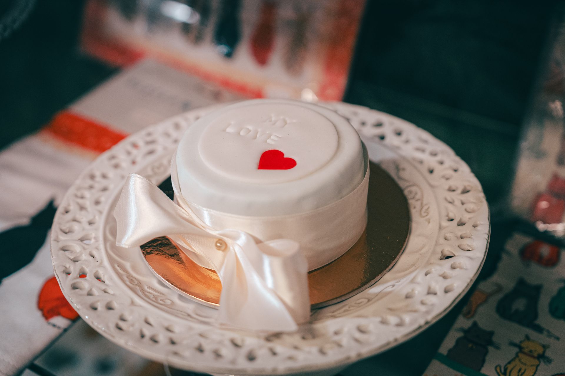 Piccola torta bianca con un nastro e un cuore rosso su un piatto bianco decorativo.