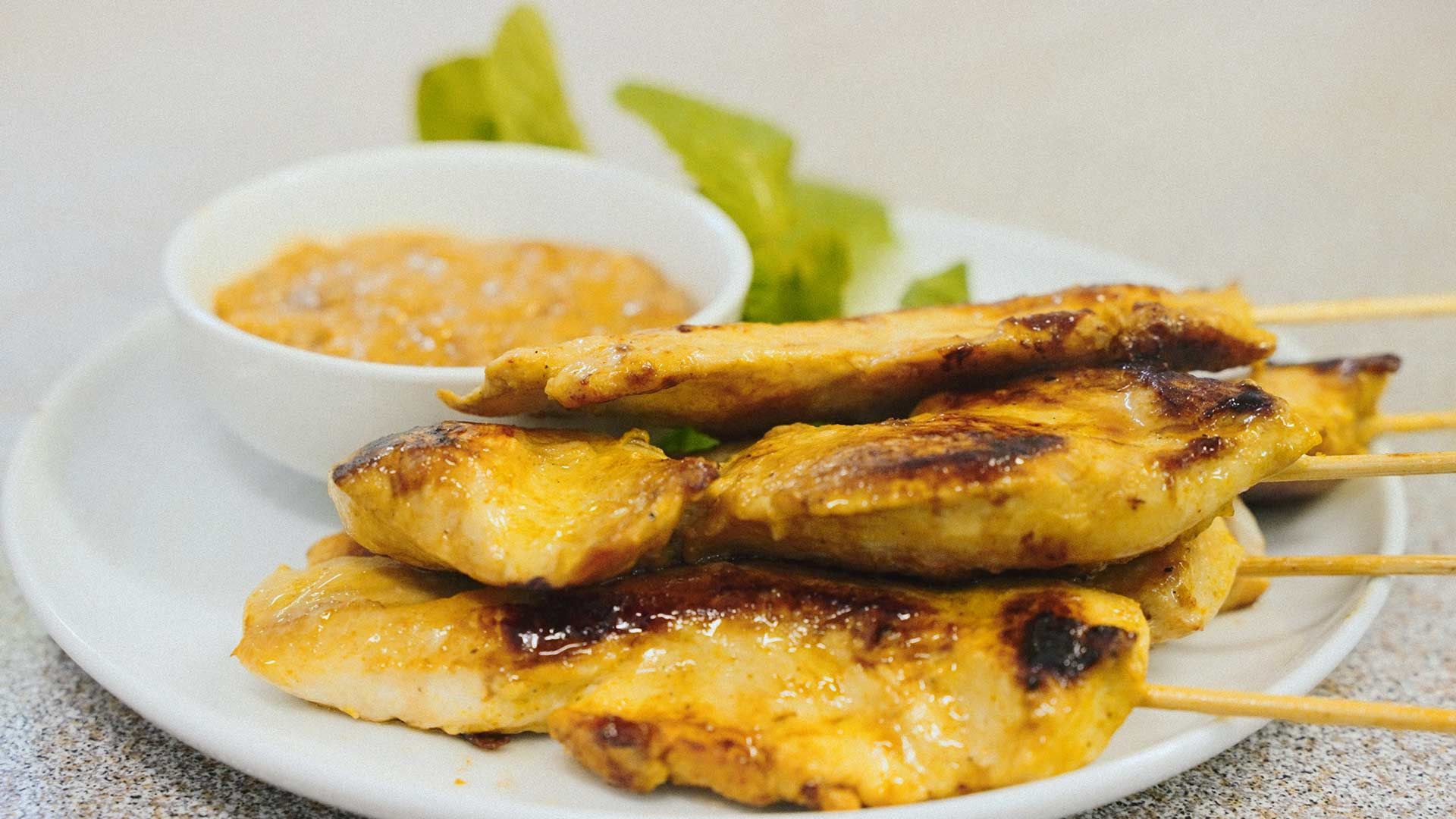 A White Plate Topped With Chicken Skewers and a Bowl of Dipping Sauce — One Thai Cuisine & Noodle Bar In Kirwan, QLD