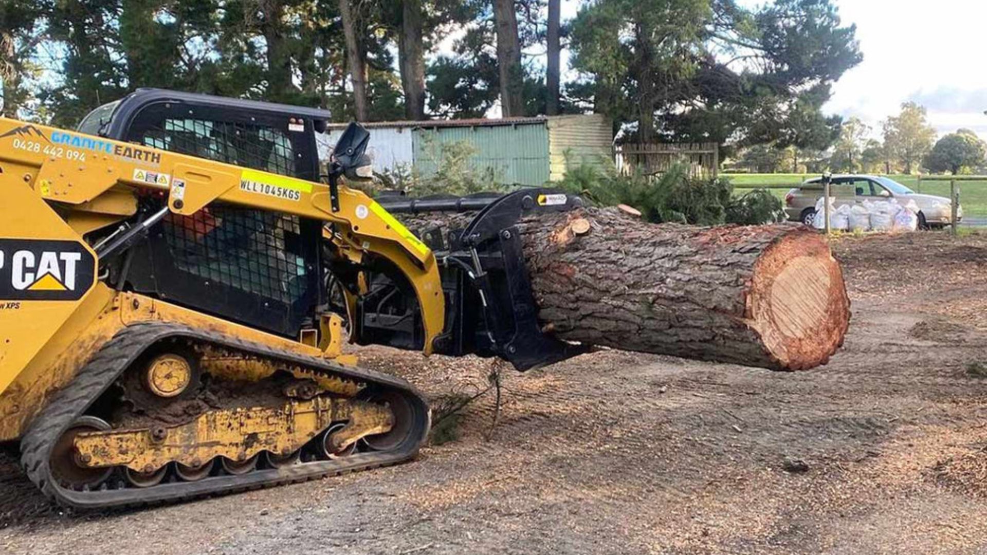 Yellow CAT skid steer pushing a large log across a dirt lot under trees