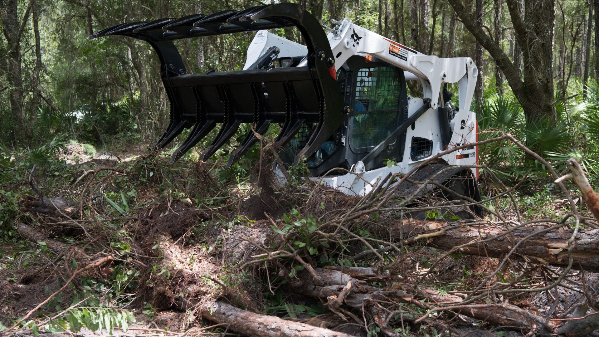 A white excavator with a black grapple sits in a wooded area amid fallen branches and brush.