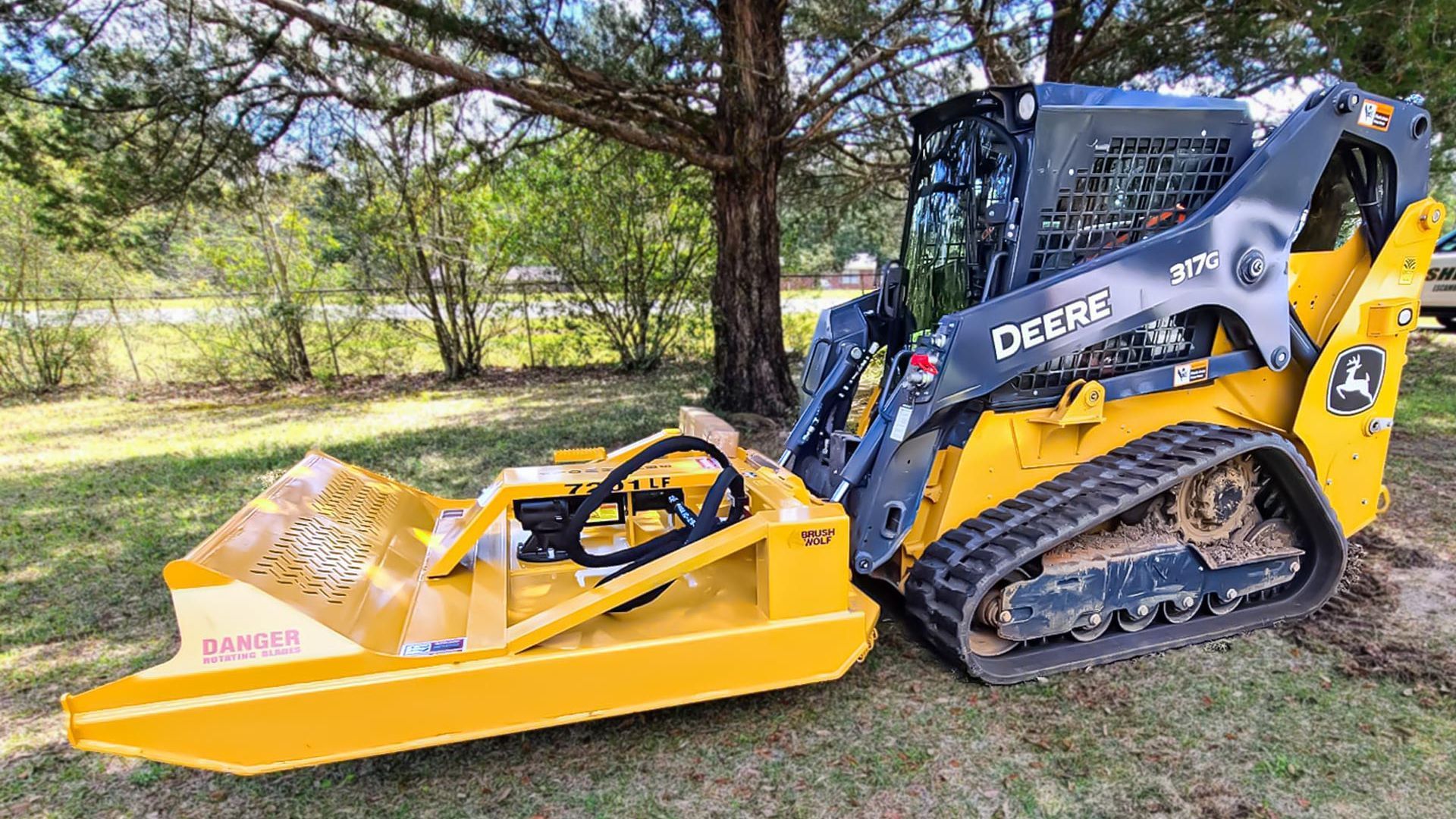 Yellow skid steer with front-mounted brush mower parked on grass under trees.