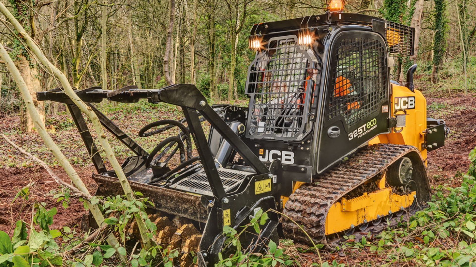 Yellow JCB skid steer mulching brush in a wooded area with fallen branches and debris