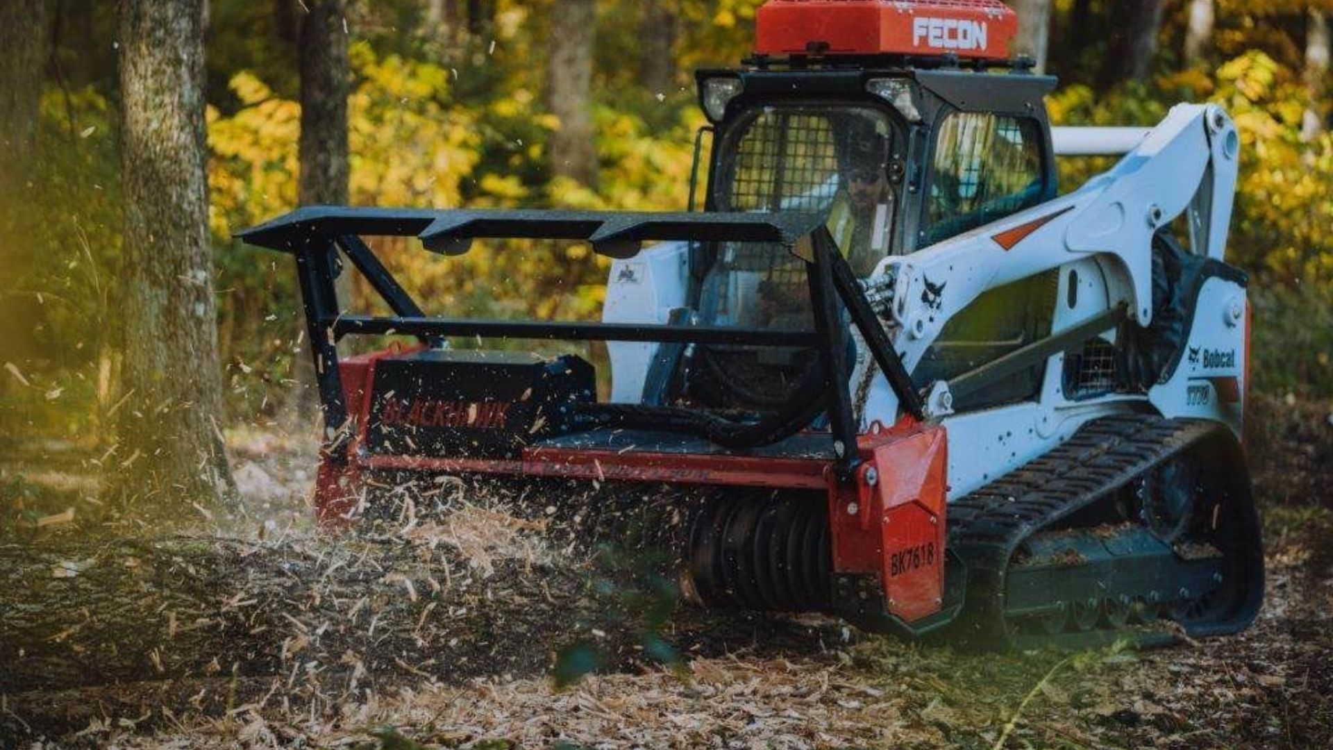 A red and white skid steer mulches brush in a wooded area, scattering leaves and debris.