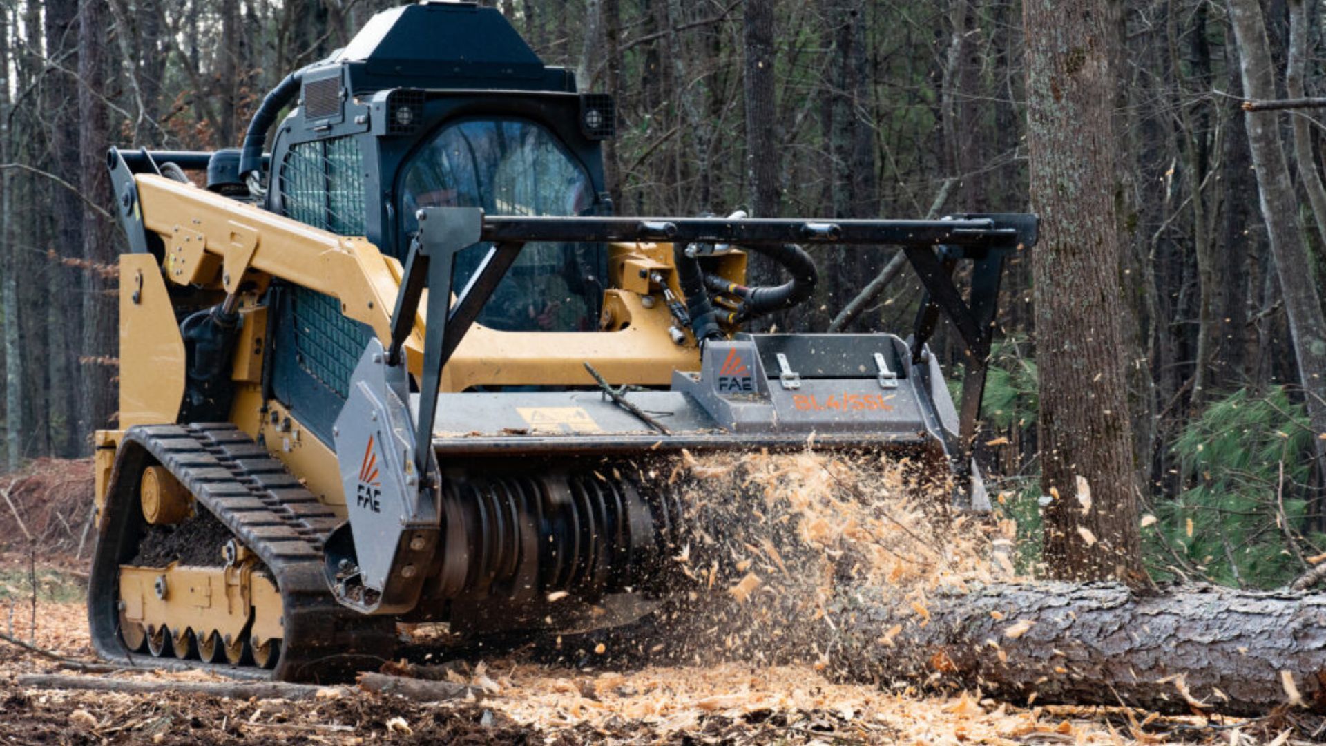 Yellow skid-steer mulcher grinding a fallen log in a wooded area, sending wood chips flying.
